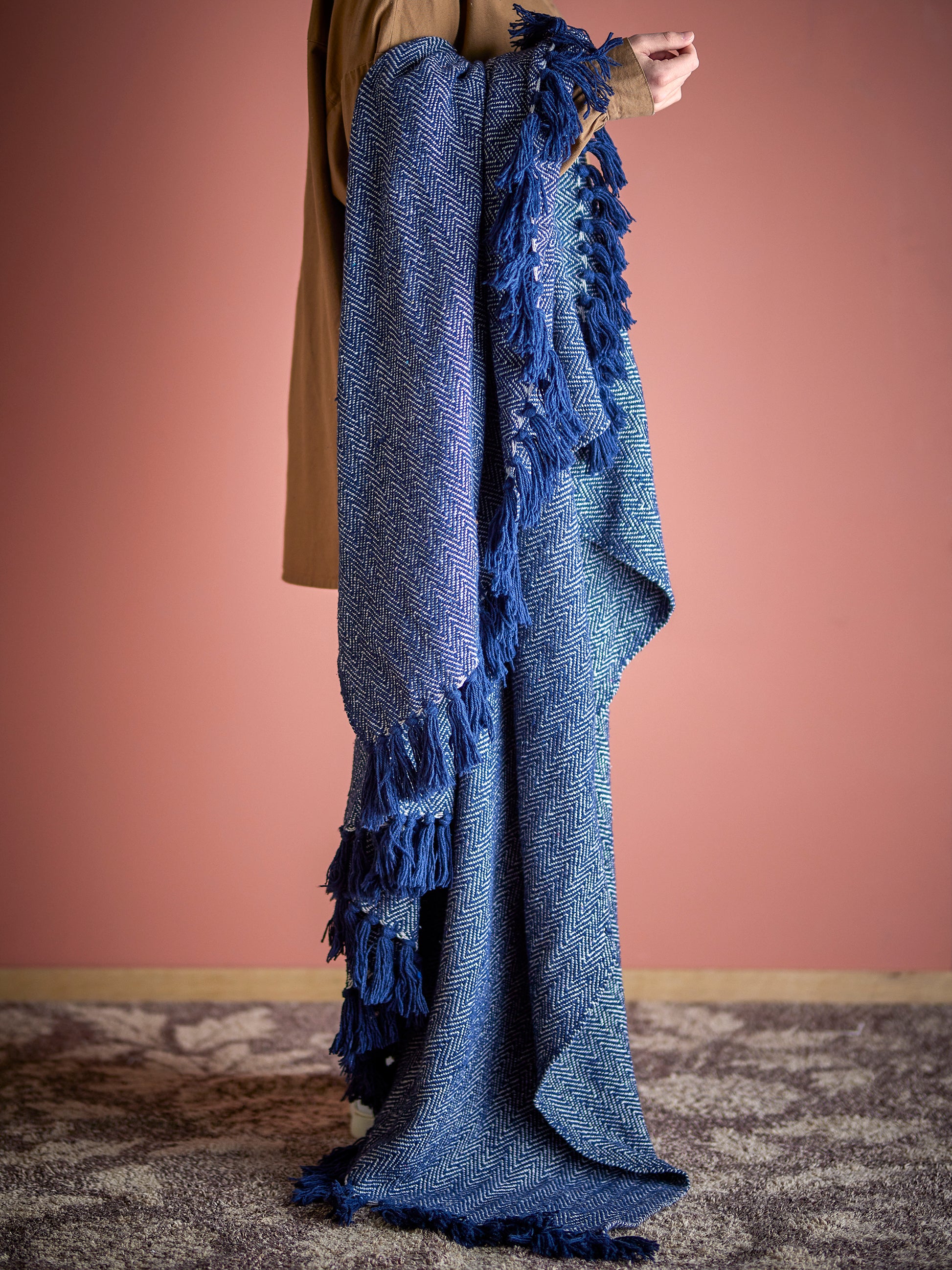 Person holding a blue knitted blanket with tassels against a pink wall.