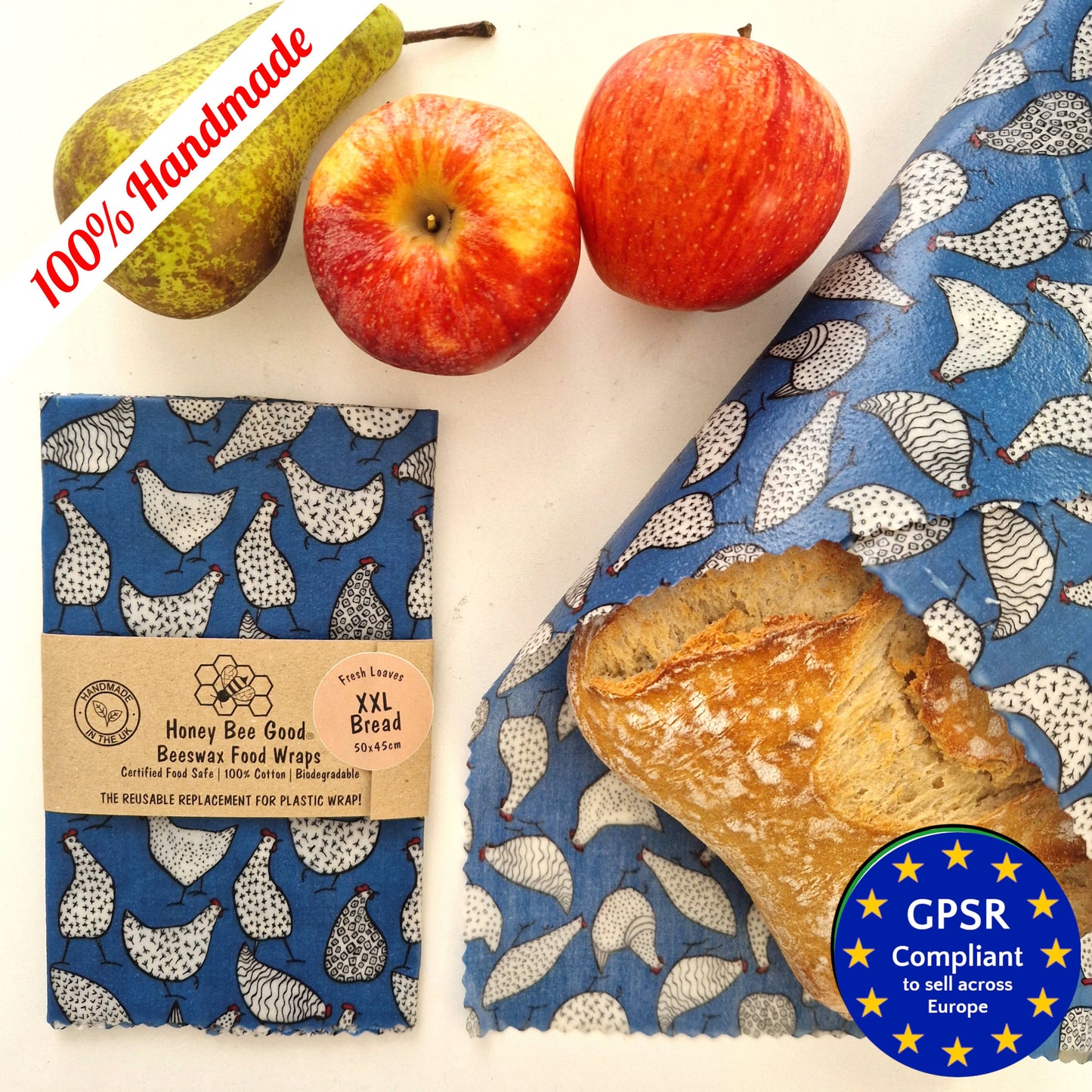 An extra large beeswax food wrap in blue with a pattern of hens, displayed next to a piece of bread, an apple, and a pear.