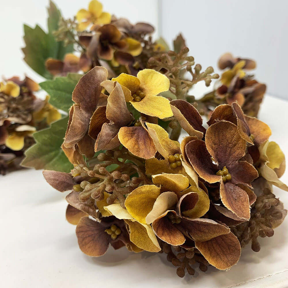 A close-up of dried or artificial hydrangea-style floral clusters with brown and tan petals, some yellow flowers, and green leaves on a plain light background.
