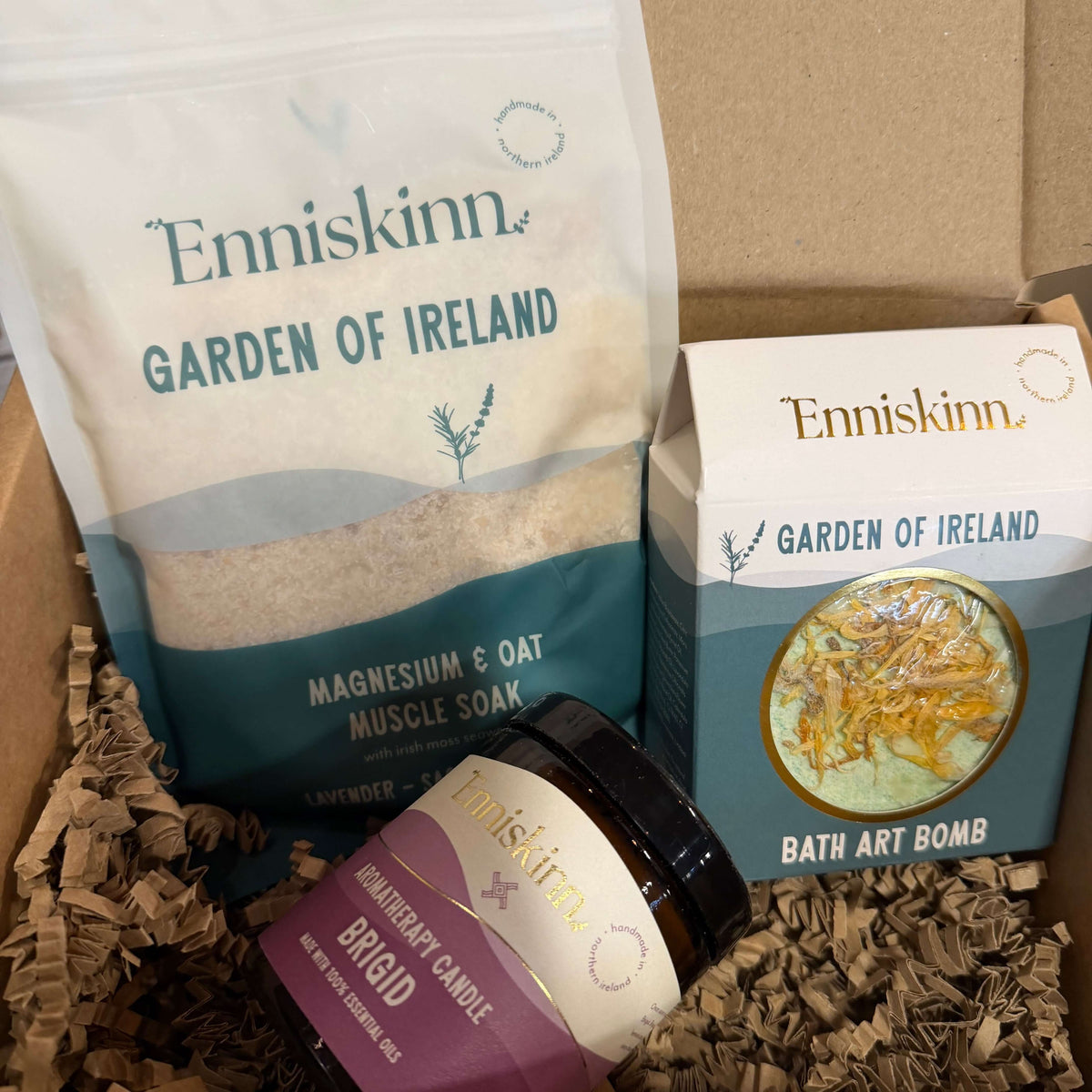An Enniskinn 'Garden of Ireland' gift box containing a large bag of Magnesium & Oat Muscle Soak, a jar of BygID Herbal Candle, and a box of Bath Art Bomb, all nestled in brown paper shred inside a cardboard box.