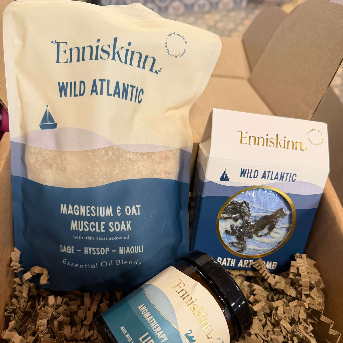 A wellness gift box from Eniskimn Wild Atlantic containing a jar of Homestudy Calf Oil, a box of Wild Atlantic, and a bag of Magnesium & Oat Muscle Soak nestled in brown crinkle paper inside a cardboard box.