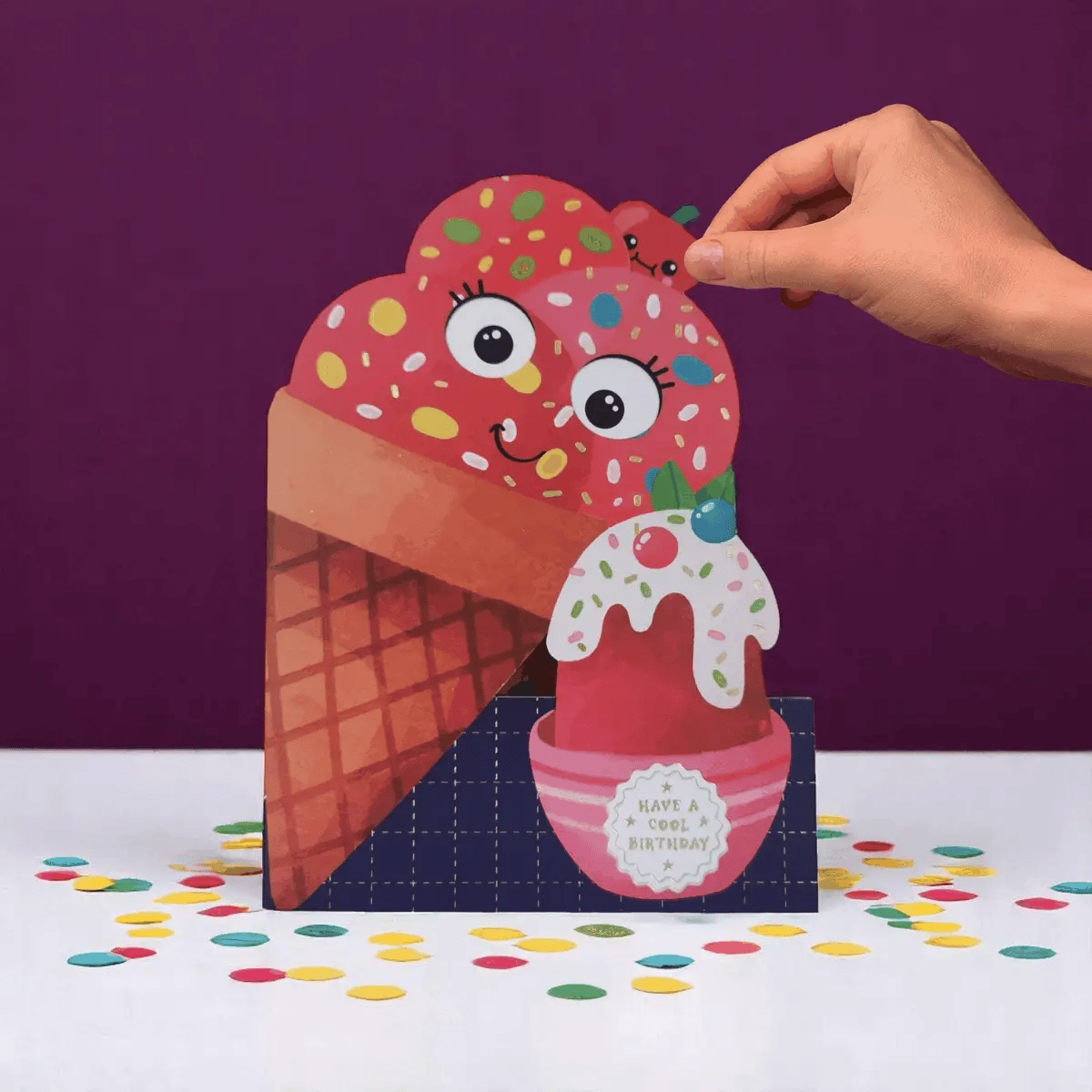Hand holding a colorful ice cream cone-shaped card with a smiling face on a purple background.