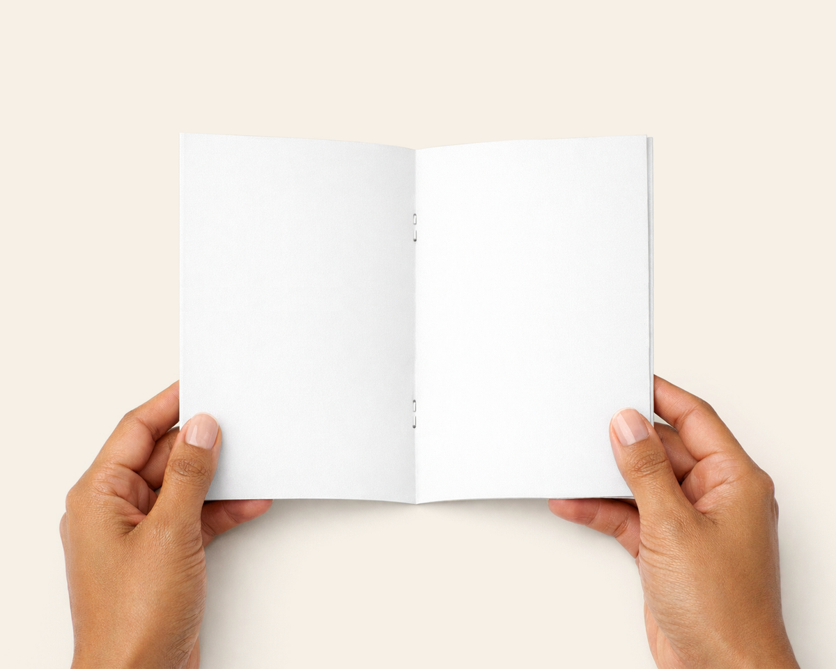 A top-down view of a person's hands holding an open, blank white booklet or notebook against a solid light beige background.