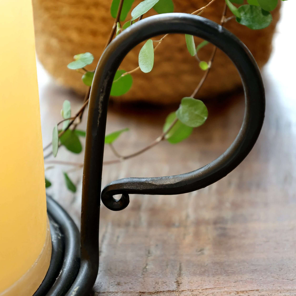A close-up of a black metal candle holder with a curved scroll design, holding a yellow pillar candle on a rustic wooden surface with a blurred plant in the background.
