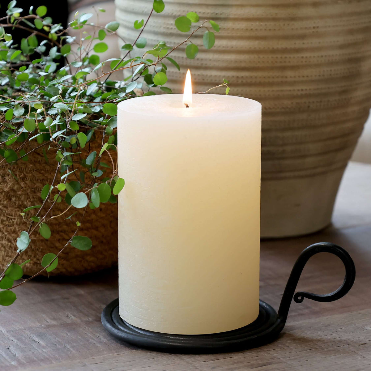 A lit cream-colored pillar candle in a black metal holder with a scroll handle, styled on a wooden surface with potted green plants in the background.