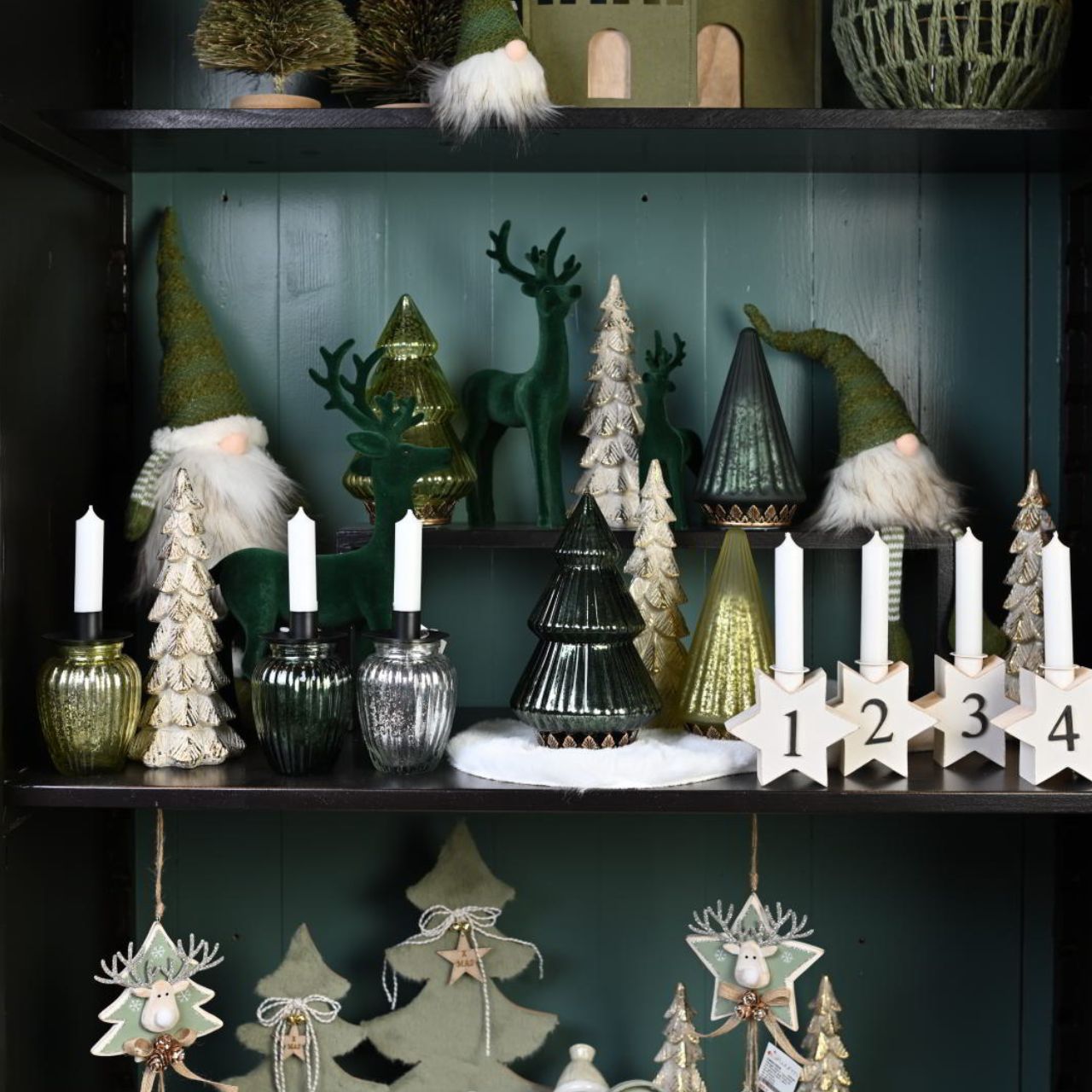 Decorative shelf with Christmas-themed items including gnomes, trees, and candles against a dark green wall.