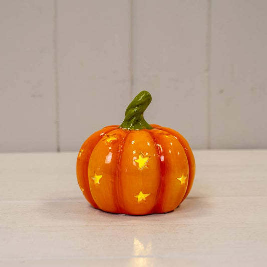 An orange ceramic pumpkin decoration with a green stem and yellow star-shaped cutouts, displayed on a white surface against a white paneled background.