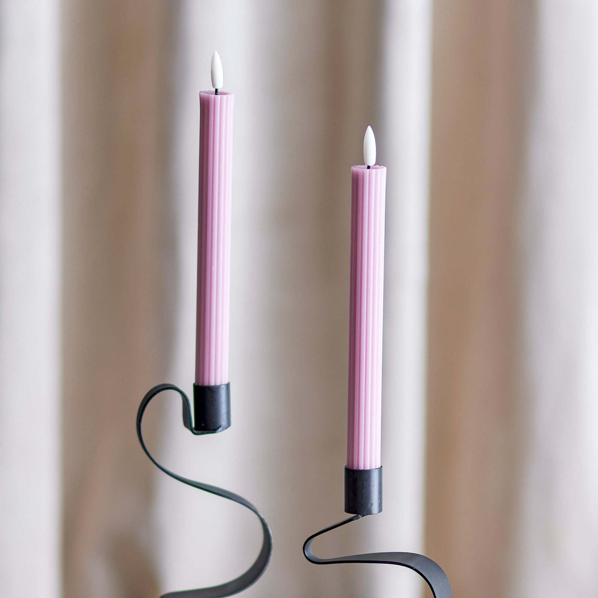 Two pink taper candles in black metal spiral candlesticks, displayed on a light-colored table against a soft beige curtain background.