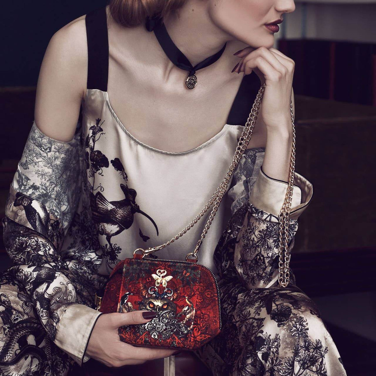 A woman in a black and beige patterned robe holds a red embroidered clutch bag with a gold chain strap. The clutch features detailed gold and silver embroidery depicting a phoenix and Chinese characters on a red background.