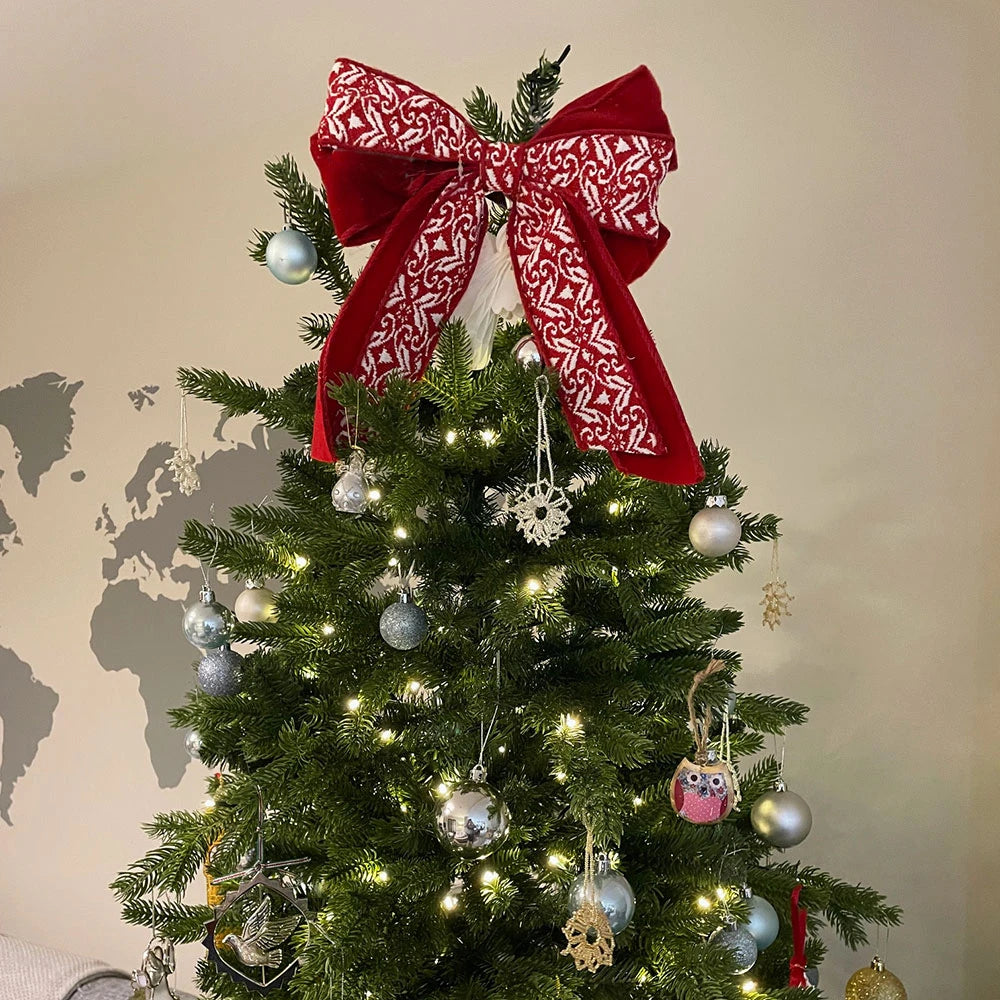 A large red fabric bow with a white patterned design, styled on top of a decorated Christmas tree with silver ornaments and warm white lights, set against a beige wall with a world map graphic.