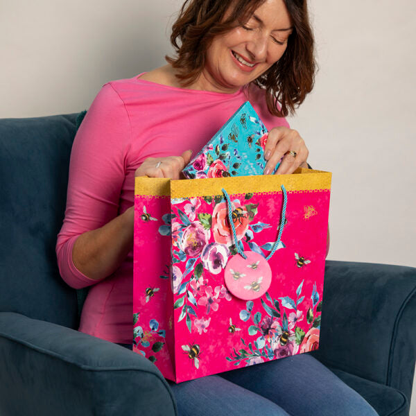 Woman opening a floral gift bag with a smaller floral box inside, sitting on a blue couch.