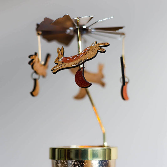 A candle carousel kit with a glass votive and a wooden carousel attachment with four laser-etched wooden ornaments of hares, displayed on a blue surface.