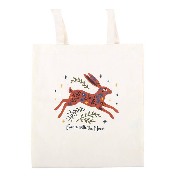 An off-white canvas tote bag featuring a whimsical illustration of a hopping rabbit in rust red and blue patterns, surrounded by green leaves, small stars, and the text 'Dance with the Moon'.
