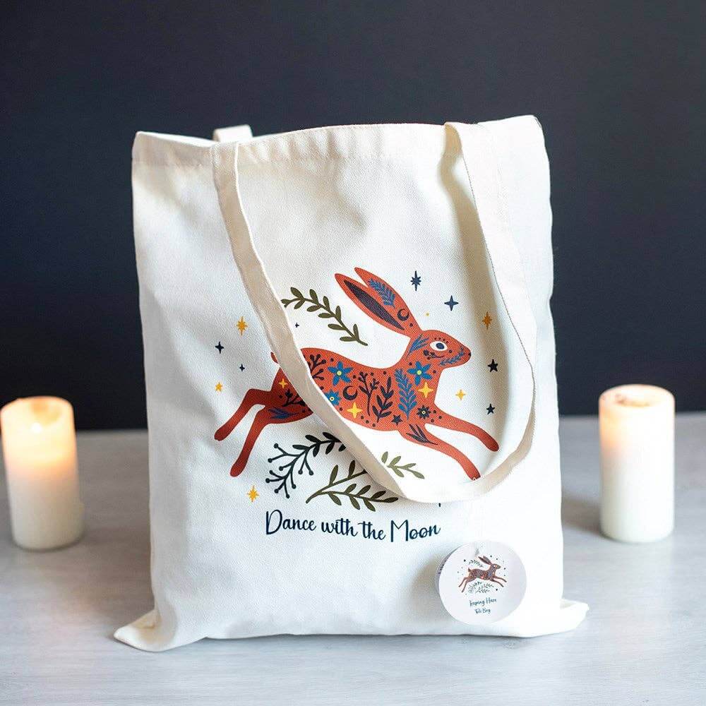 A cream canvas tote bag with a colorful illustration of a hopping hare and the text 'Dance with the Moon', styled on a tabletop with two lit candles in the background.
