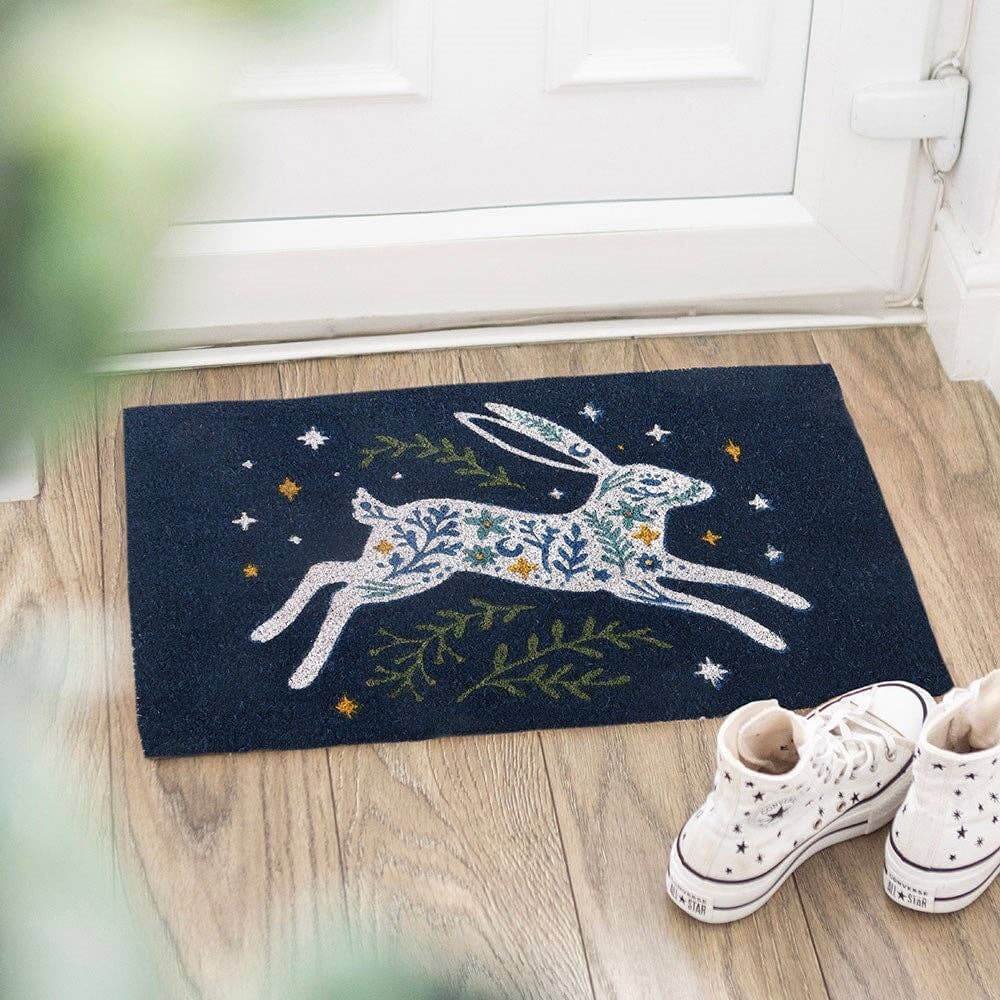 Decorative doormat with a hare design on a wooden floor, next to white sneakers.
