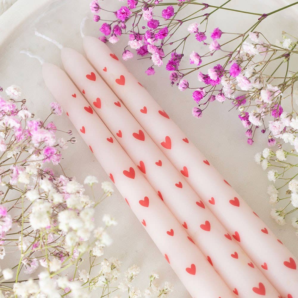 Pink taper candles with red heart patterns on a white surface with pink and white flowers.