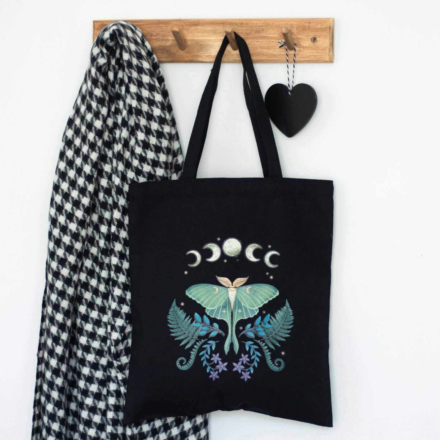 Black tote bag with celestial design hanging on a wooden hook with a checkered scarf and black heart.