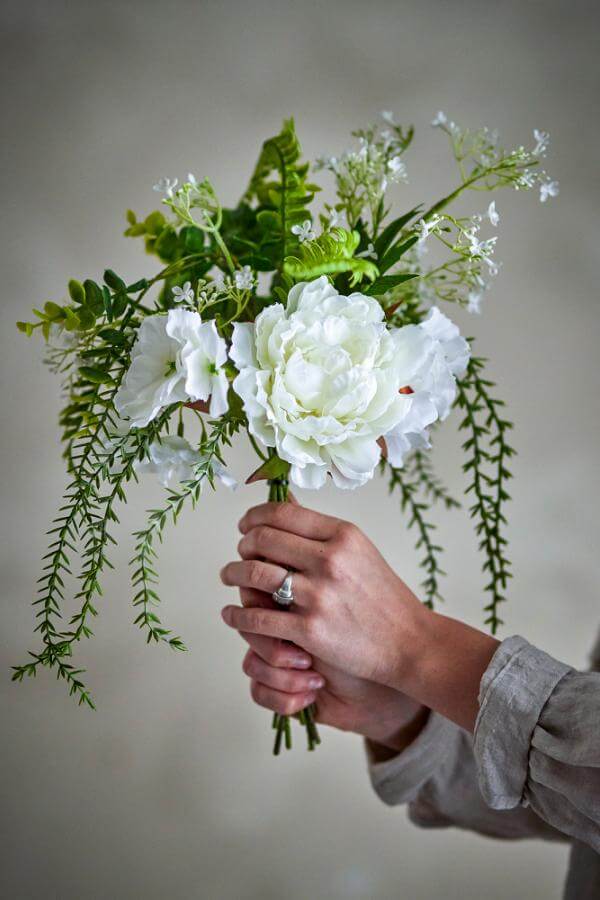 Hand holding a bouquet of white flowers and green leaves against a neutral background
