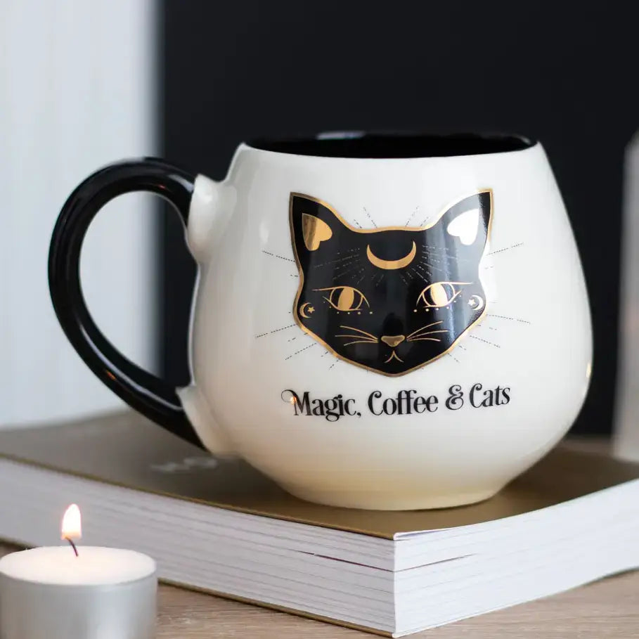 A white ceramic mug with a black handle and black interior, featuring a black cat face with gold details and the text 'Magic, Coffee & Cats' on the front. The mug sits on a book with a lit candle in the foreground.