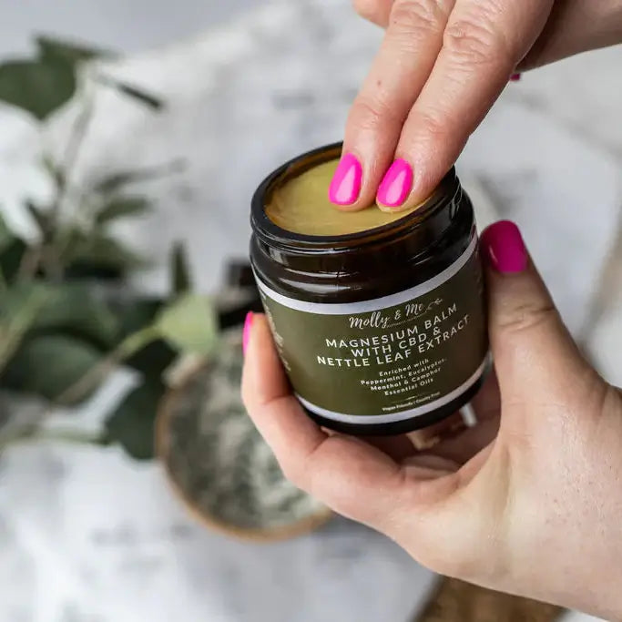 Hand holding a jar of magnesium balm with a blurred plant in the background