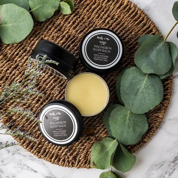 Three containers of magnesium sleep balm on a woven mat with green leaves around.