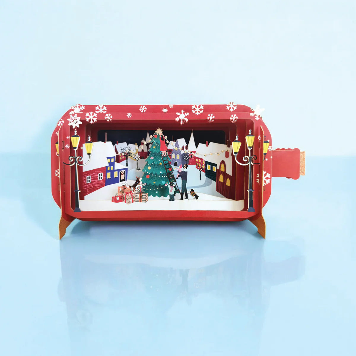 Pop-up Christmas card showing family decorating a Christmas tree in a snowy town centre with dogs, presents, and a festive 3D scene.