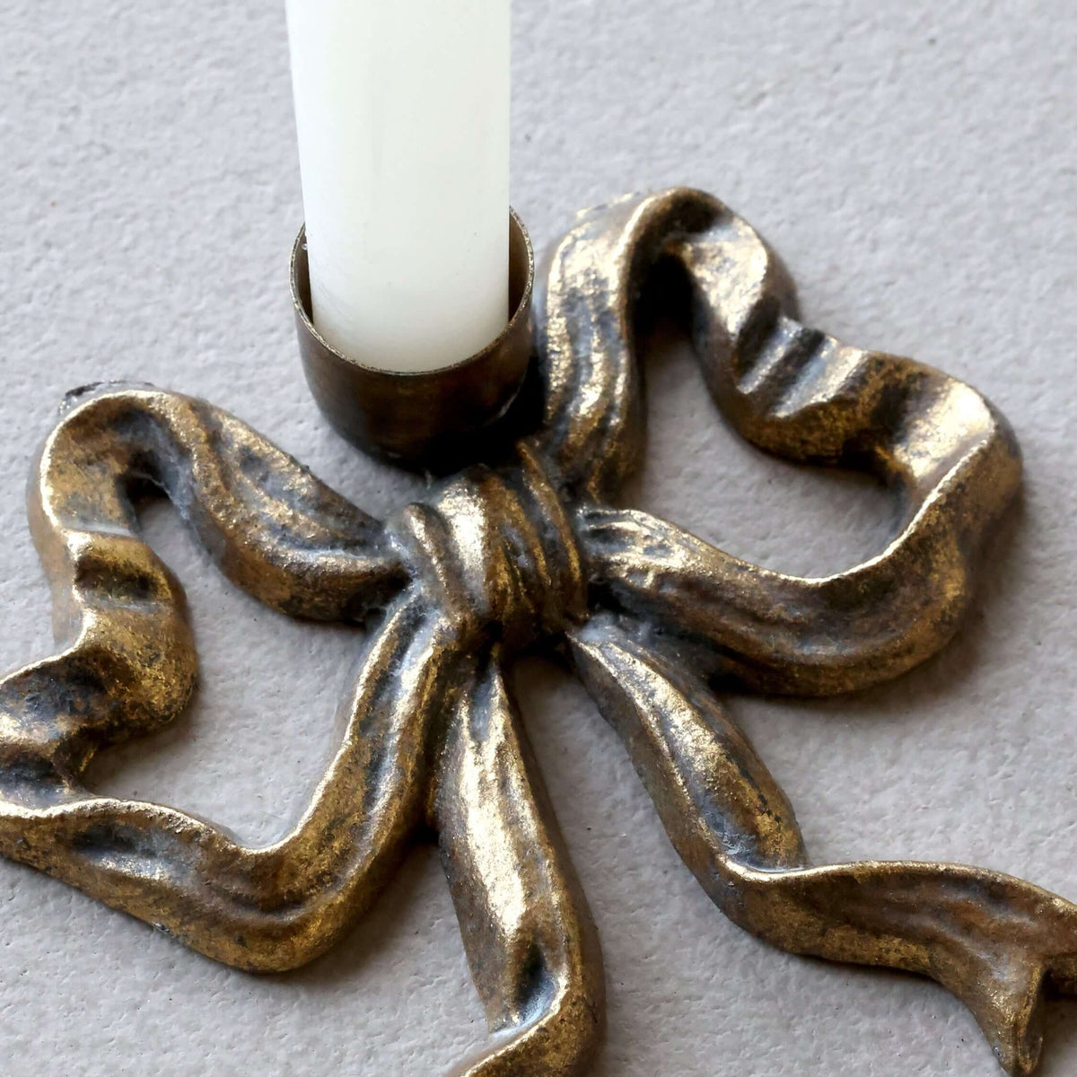 A close-up of a metallic bow-shaped candle holder with a white candle inserted, set against a neutral gray background.