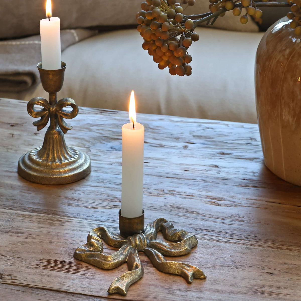 Two lit white pillar candles in ornate metallic holders on a rustic wooden table, with a beige sofa and decorative vase in the softly blurred background.