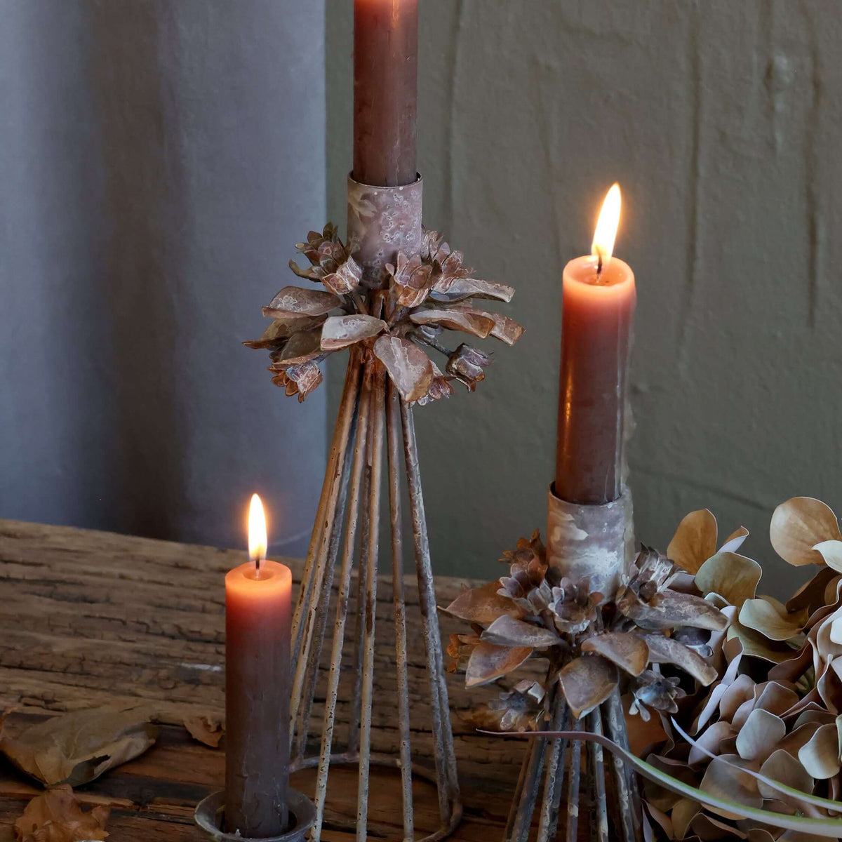 Three rustic brown taper candles in decorative metal holders, including a shell-shaped base, a wire cone with pinecone accents, and a wire cone with dried flowers, arranged on a wooden surface with scattered autumn leaves.