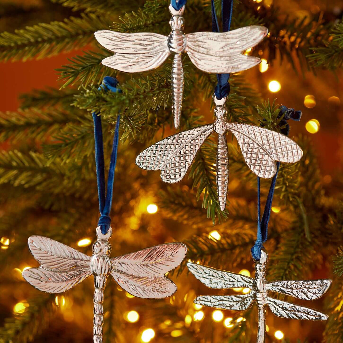 Four metallic dragonfly Christmas ornaments in silver finish hanging from a green Christmas tree with warm fairy lights in the background.