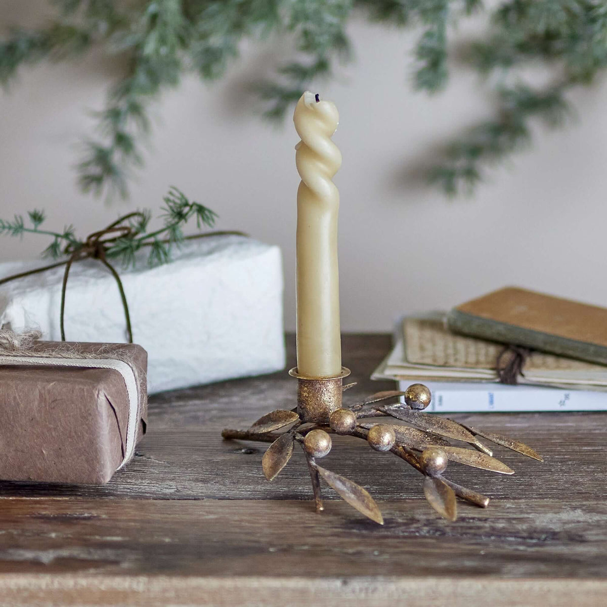 A twisted cream candle in a decorative gold-toned metal holder with leaf and bead details, styled on a rustic wooden table with wrapped gifts and books in the background.