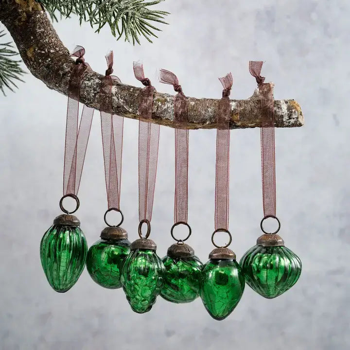 Five green ribbed glass Christmas ornaments hang from a tree branch with sheer mauve ribbons against a soft gray background.