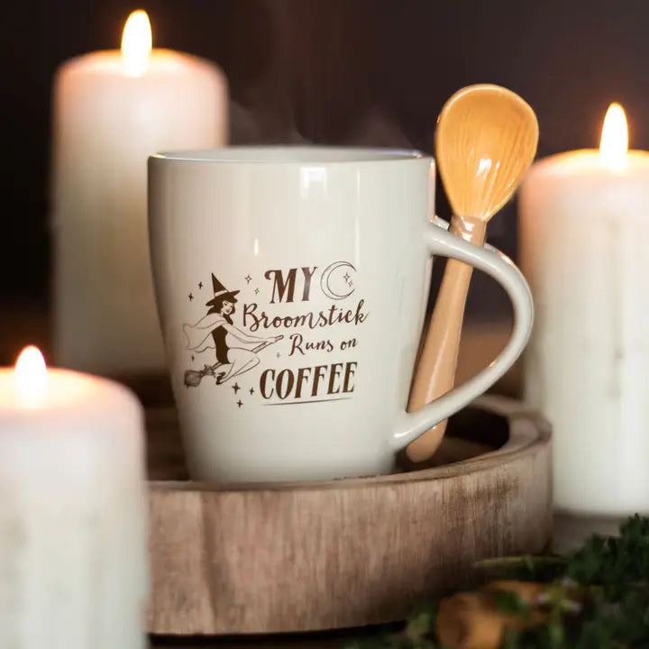 White mug with 'My Broomstick Runs on Coffee' text, wooden spoon, and lit candles in the background