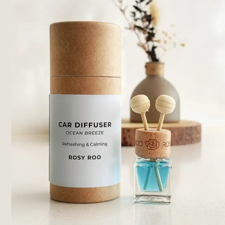 A small glass reed diffuser bottle with a light wood cap and two wooden diffuser sticks, placed on a white surface in front of a ceramic vase with dried stems on a round wood slice.