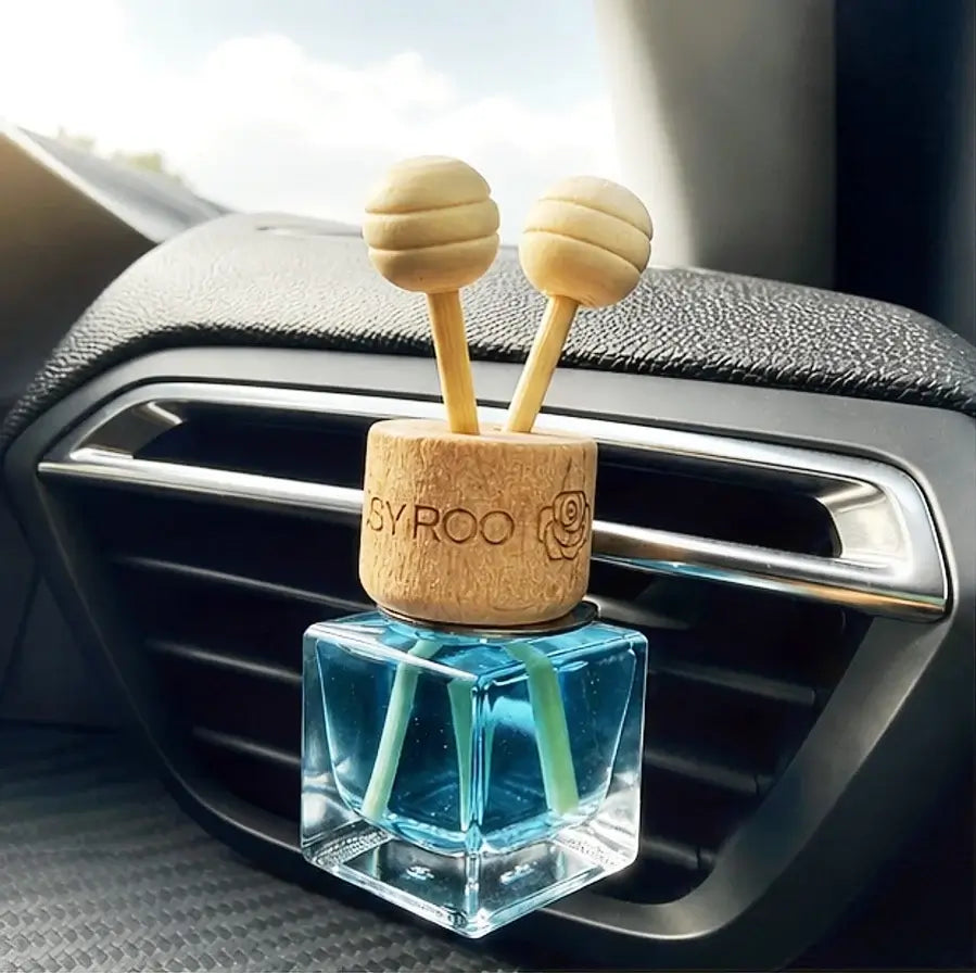 A car air freshener with a clear square glass bottle filled with blue liquid and two light green reeds, clipped onto a car air vent. The top cap is made of natural wood with two spherical wooden diffuser heads sticking out.