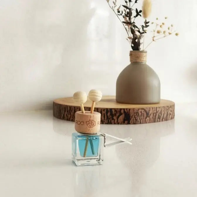 A small reed diffuser bottle with blue liquid and wooden diffuser sticks sits on a glossy white surface, styled in front of a ceramic vase with dried stems on a wood slice tray.