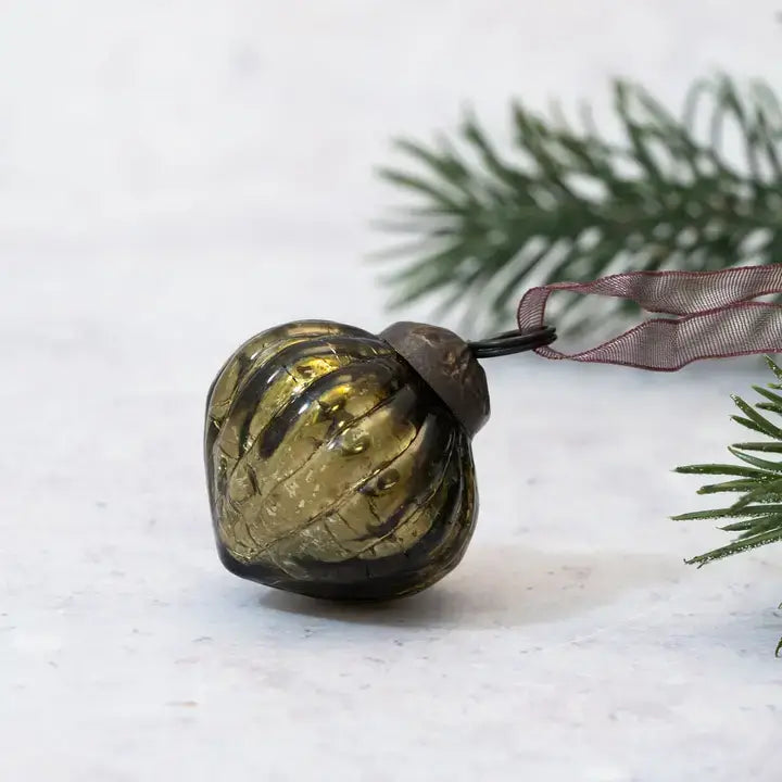 A vintage-style glass Christmas ornament with a ribbed, bulbous shape in a smoky olive-brown and gold color, featuring a dark metal cap and a sheer mauve ribbon. The ornament is styled on a light marble surface with blurred green pine needles in the background.