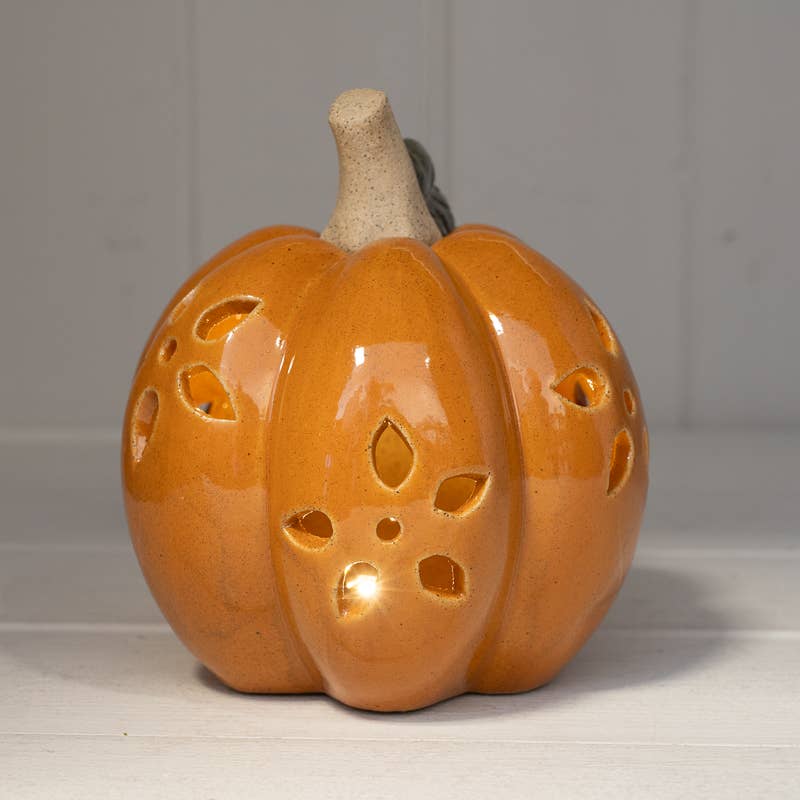 An orange ceramic pumpkin-shaped lamp with cutout holes on the front and sides, showing a warm light glowing from within, set against a neutral gray background.