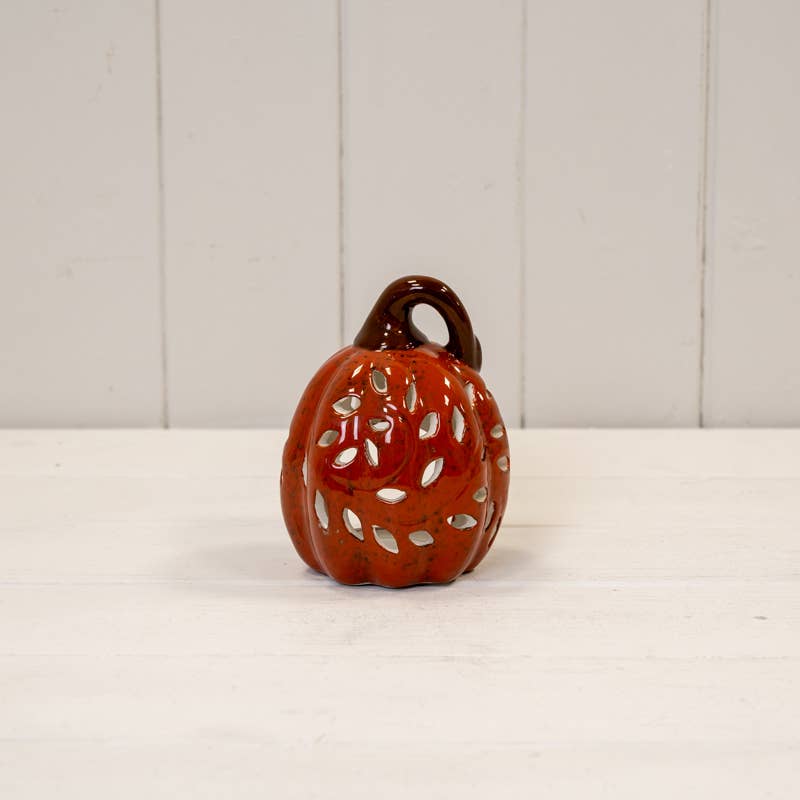 A small decorative ceramic pumpkin with a glossy orange-red glaze, featuring cutout leaf patterns and a dark brown stem, set on a white surface against a white paneled background.