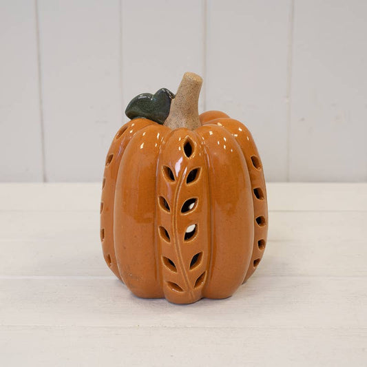 An orange ceramic pumpkin-shaped candle holder with leaf-shaped cutouts, a beige stem, and a small dark green leaf on top, set against a white paneled background.
