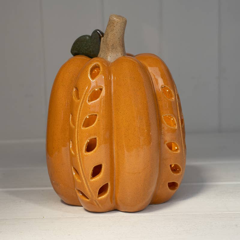 A glazed ceramic pumpkin figurine with leaf-shaped cutouts, featuring a warm orange body, a light tan stem, and a small dark green leaf on top, set against a light gray paneled background.