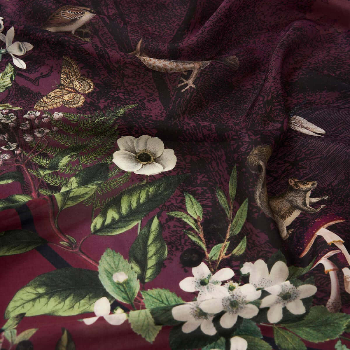 A close-up of a dark burgundy fabric with an all-over botanical and wildlife print featuring green leaves, white flowers, butterflies, birds, and small mammals.