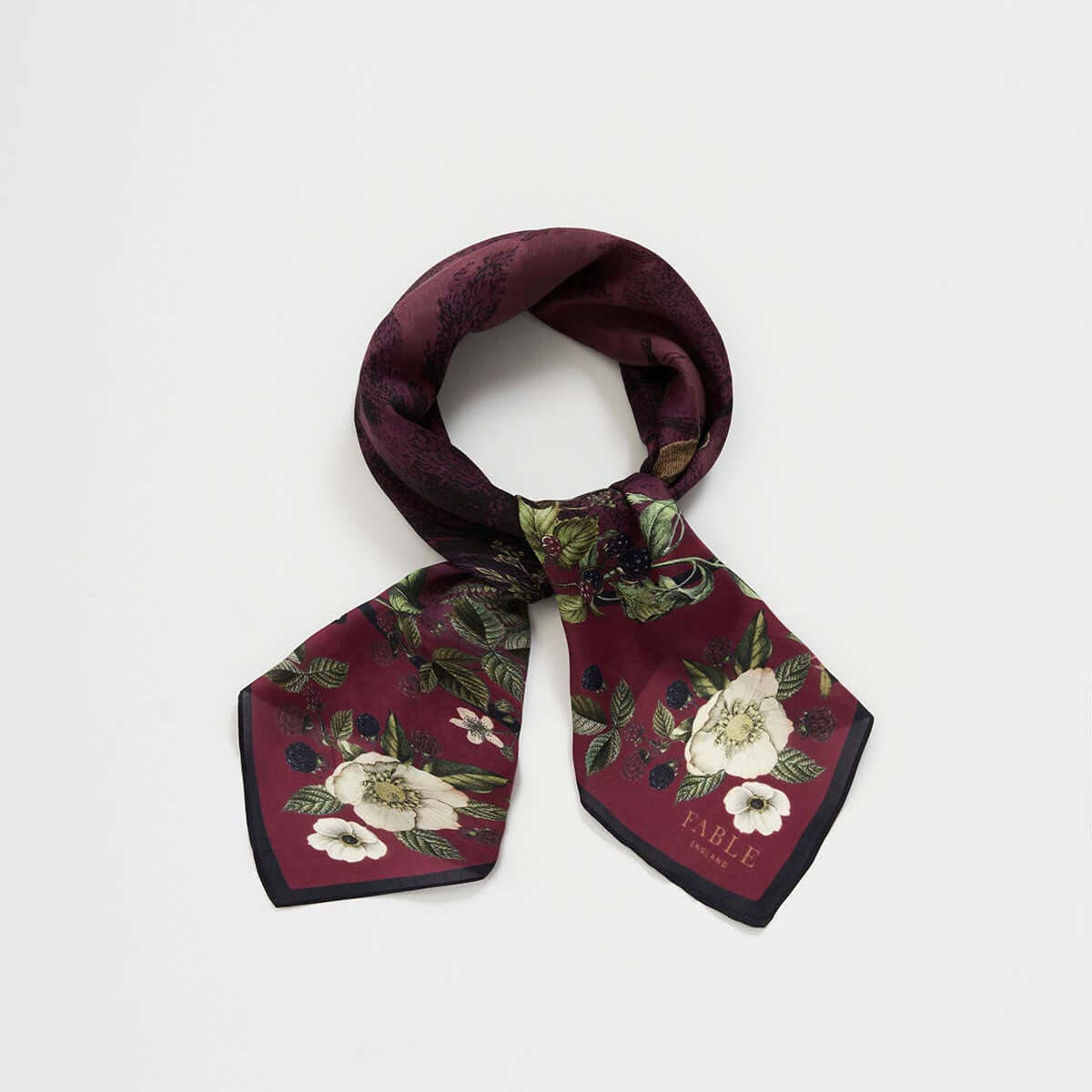 A burgundy silk scarf with a floral print featuring white flowers and green leaves, tied in a loop on a light gray background.