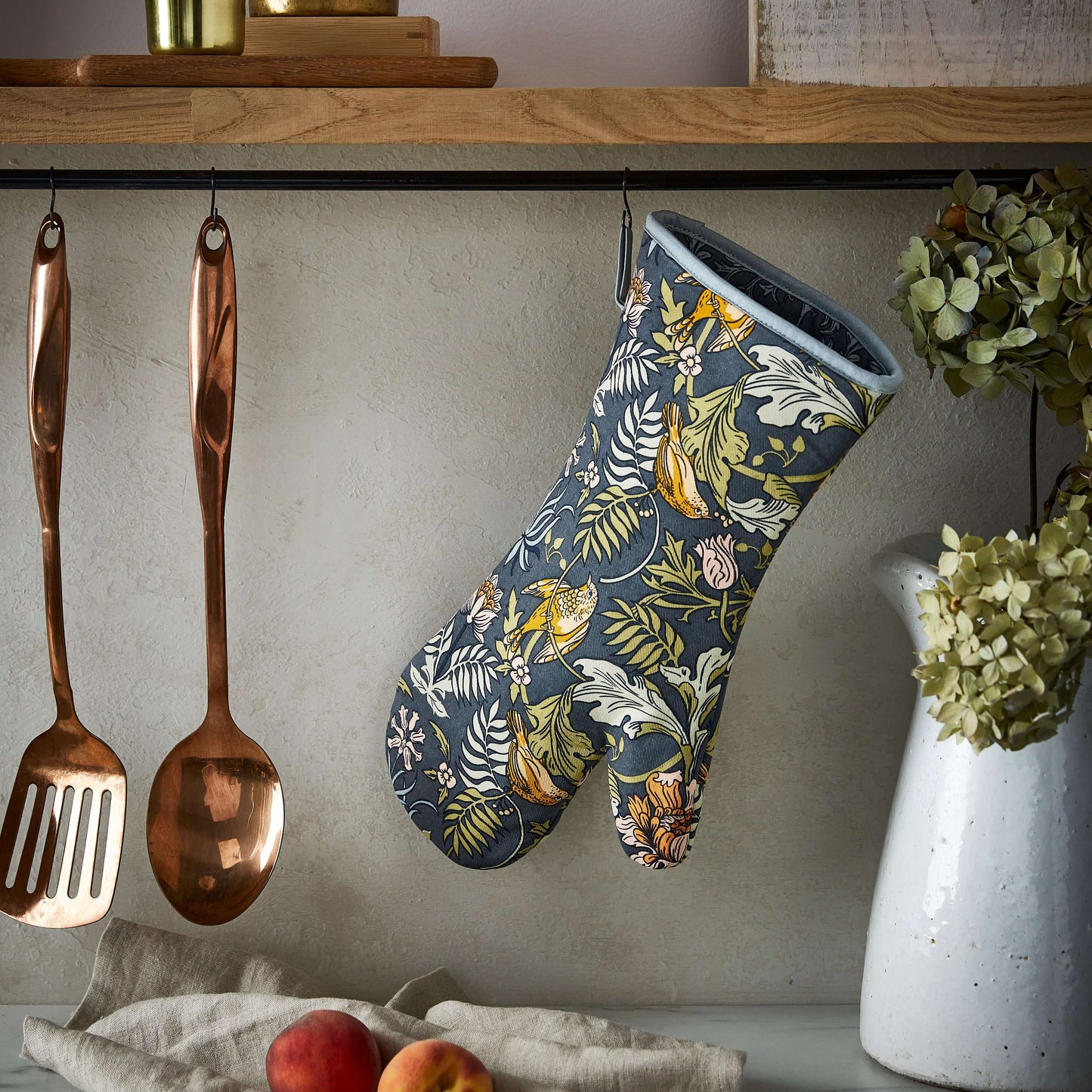 Floral-patterned oven mitt hanging on a kitchen wall with copper utensils and a vase.