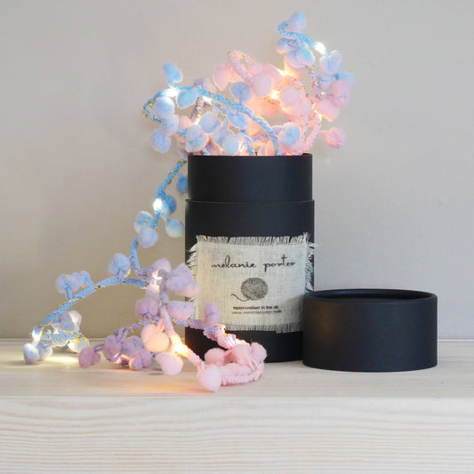 A string of fairy lights with soft white pompoms, displayed alongside a black cylindrical container with a label, against a striped background.