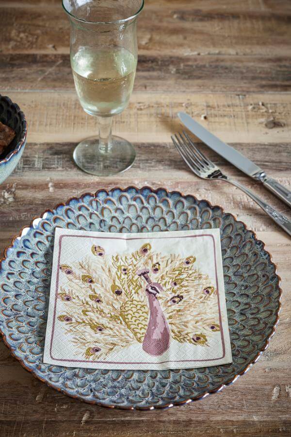 Napkin with peacock design on a textured plate with a glass of white wine and cutlery on a wooden table.