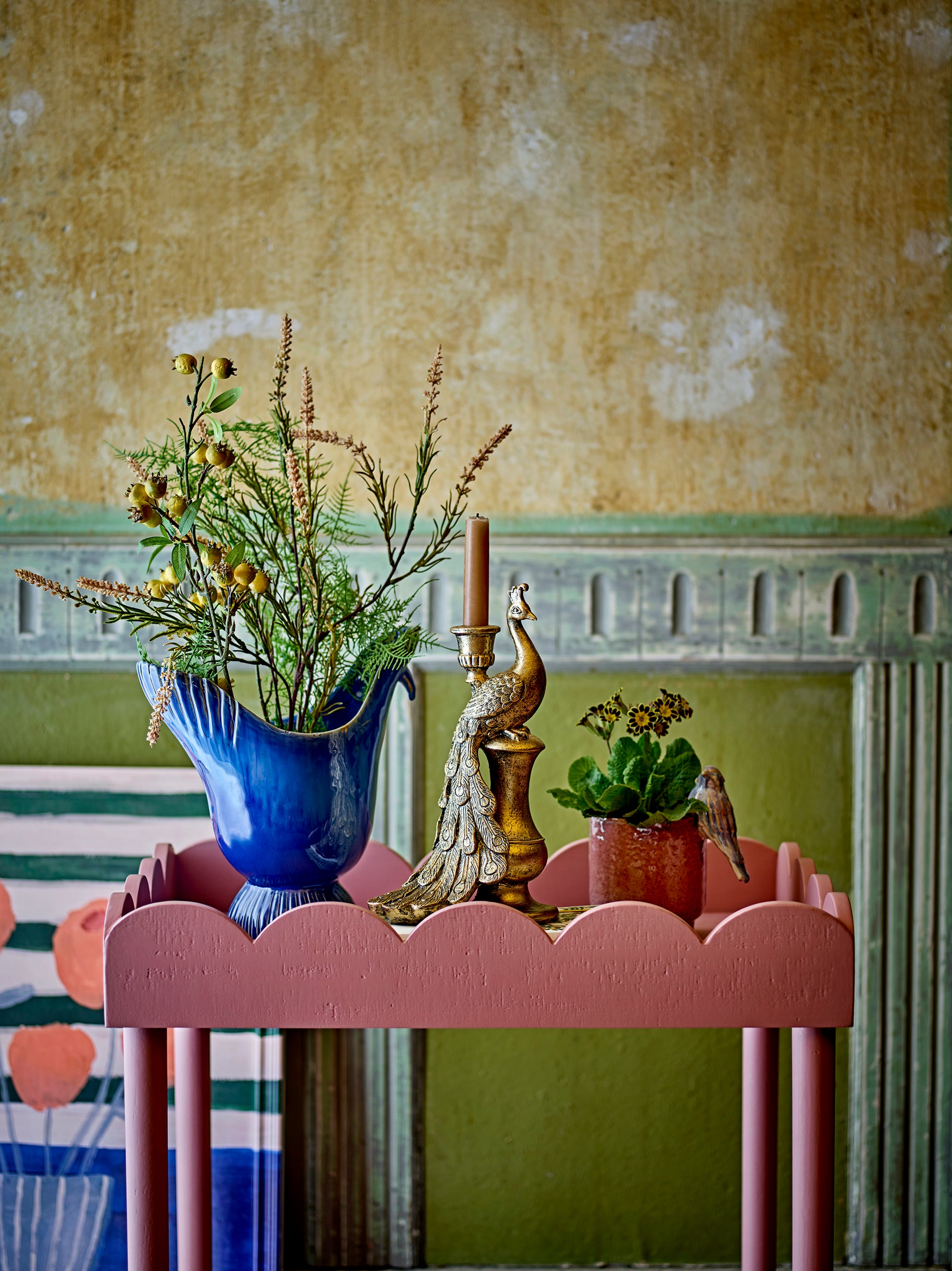 Decorative table with plants and a peacock figurine against a textured wall.