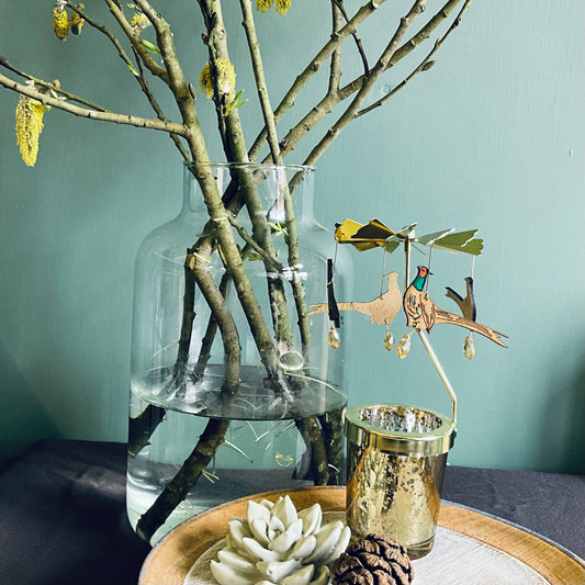 A glass votive candle holder with a gold and multicolored pheasant carousel attachment, featuring wooden ornaments in a gold vase, with a teal background and decorative items.