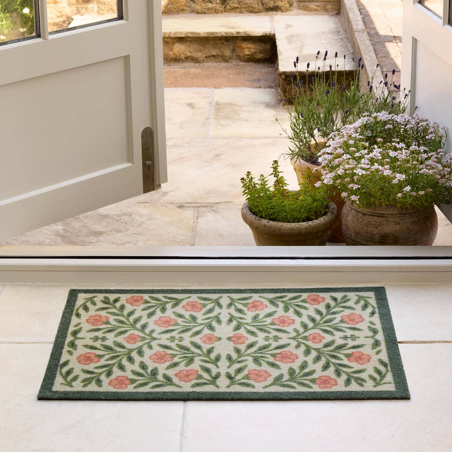 Decorative doormat with floral pattern on a white floor, leading to a door with plants outside.