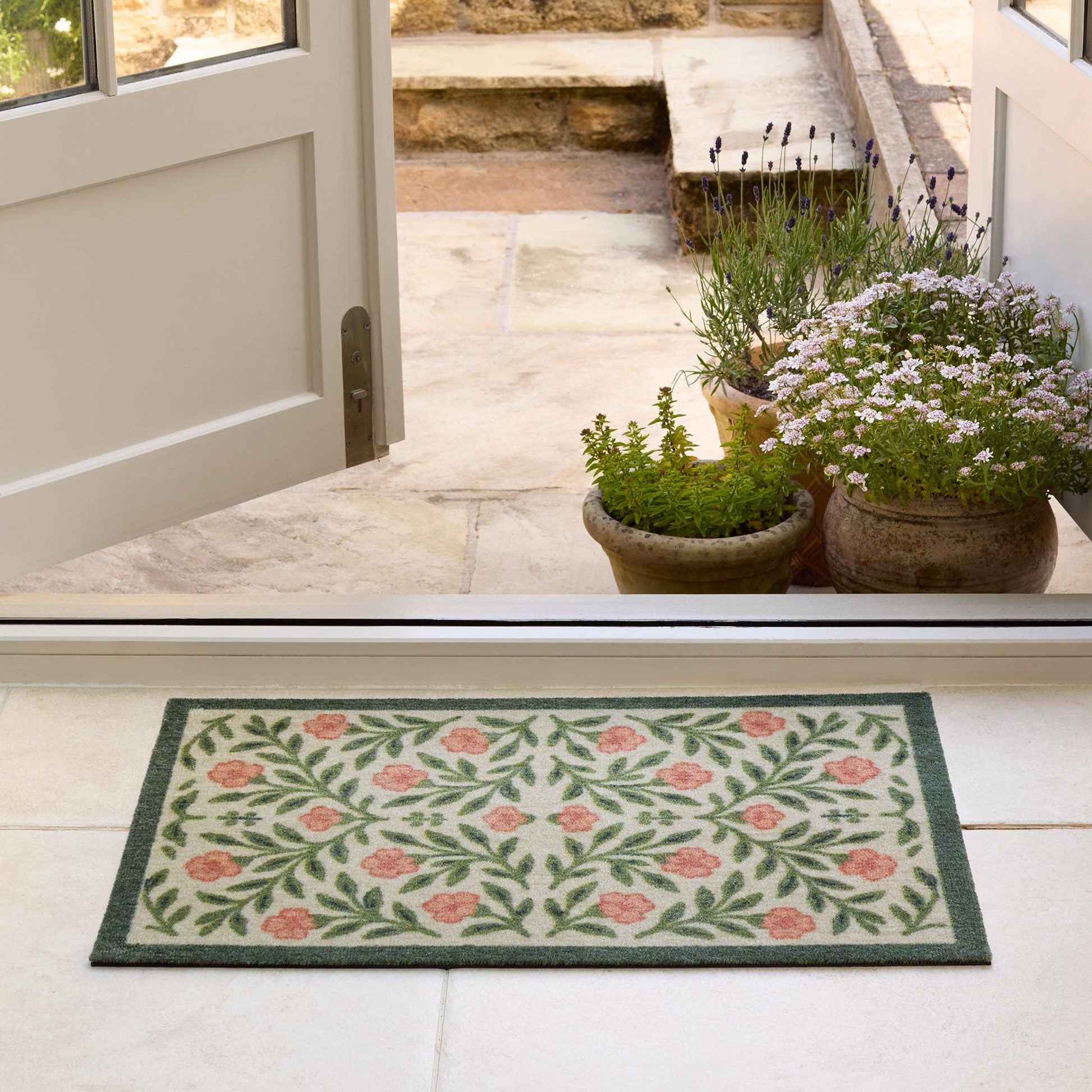 Decorative doormat with floral pattern on a white floor, leading to a door with plants outside.